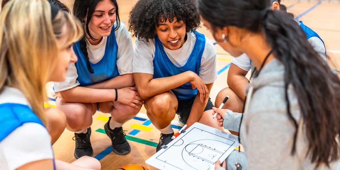 Basketball coach explaining game strategy to her young female athletes on a clipboard in a gym